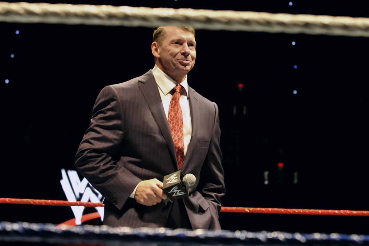 Vince McMahon wearing a suit and red tie while holding a microphone standing in a wrestling ring