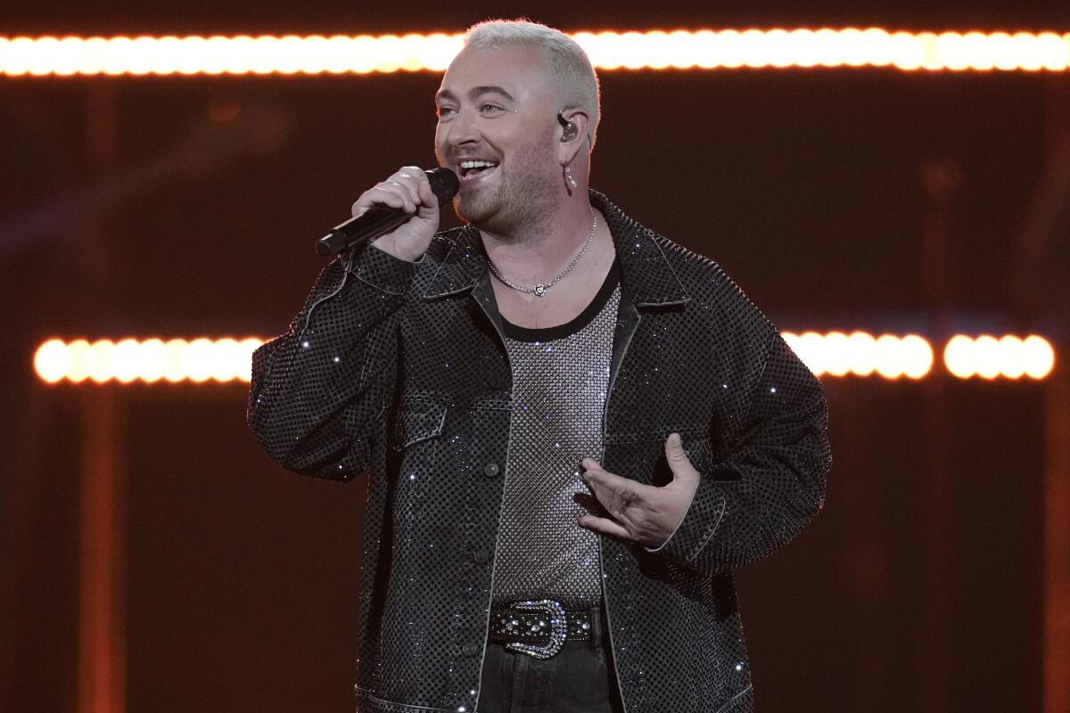 A person with short white hair singing into a microphone and wearing a sparkly jacket
