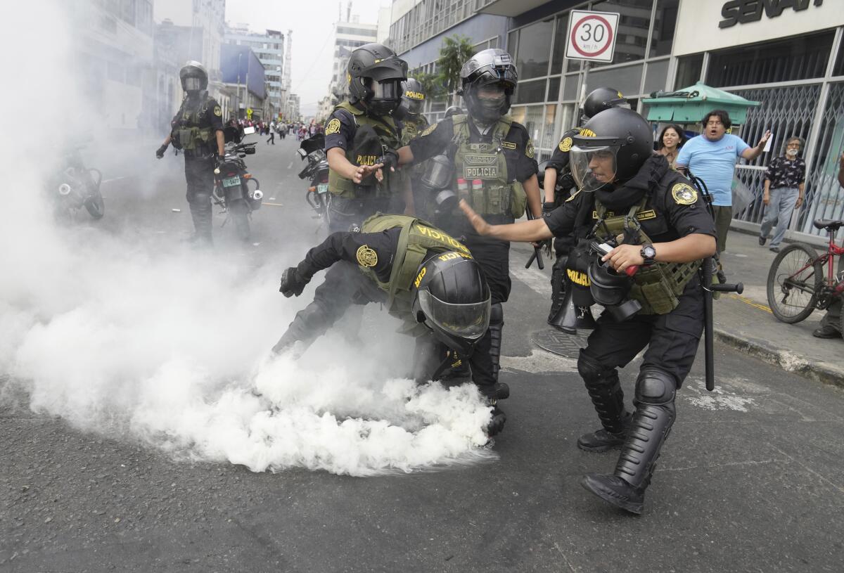 Police officers in riot gear, including one picking up a billowing tear gas canister