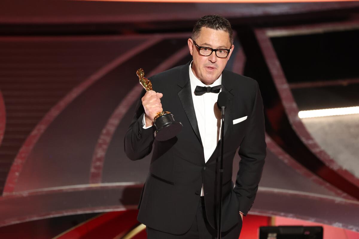 A bespectacled man wearing a tux holds a statuette on the Oscar stage.