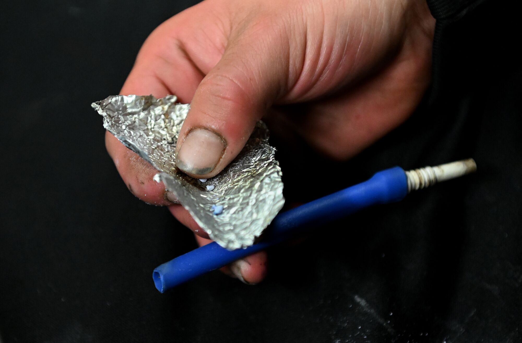 A hand with dirty fingernails holds tinfoil with blue bits of fentanyl and a makeshift plastic-tube pipe.