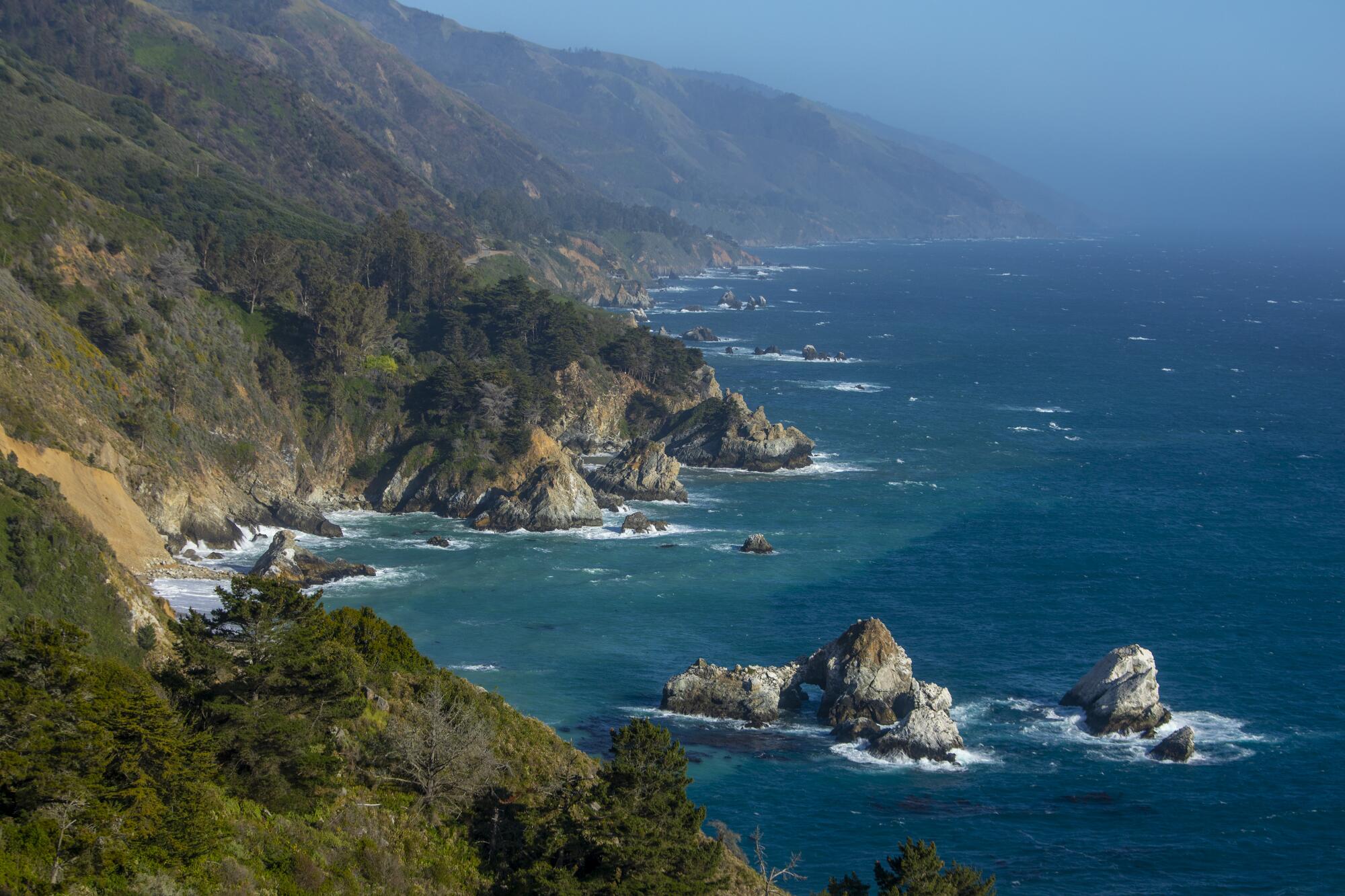 A long stretch of rocky, tree-covered coastline is met by blue-green waters and misty blue sky.  