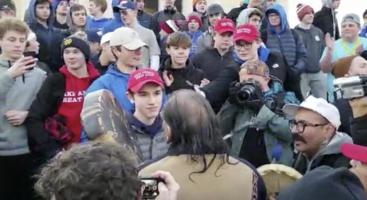 In an image from a video, a teenager wearing a "Make America Great Again" hat, center left, stands in front of an elderly Native American singing and playing a drum in Washington.