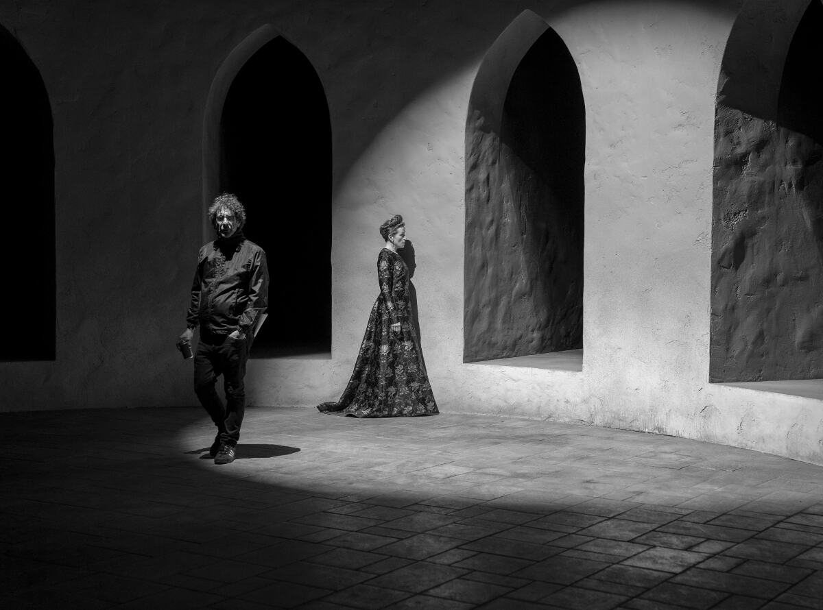 A black-and-white photo of a man walking away from a woman in a gown in front of a few arches 