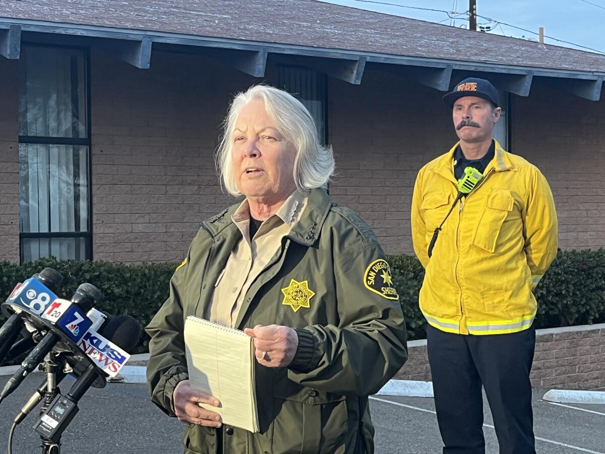 A woman with a gray bob and wearing a law enforcement uniform speaks into microphones.