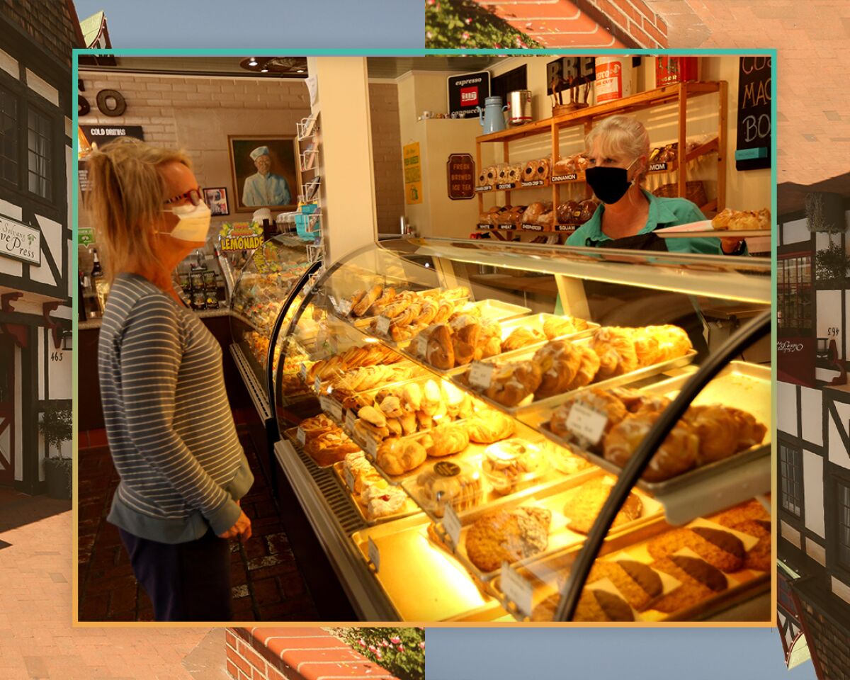 A woman in a mask stands behind a glass-front bakery case. A customer in a mask looks at the goods.