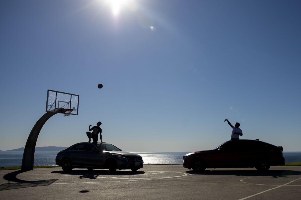 At an oceanside basketball court, a person sits on a car. A person shoots a basketball from the open sunroof of another.