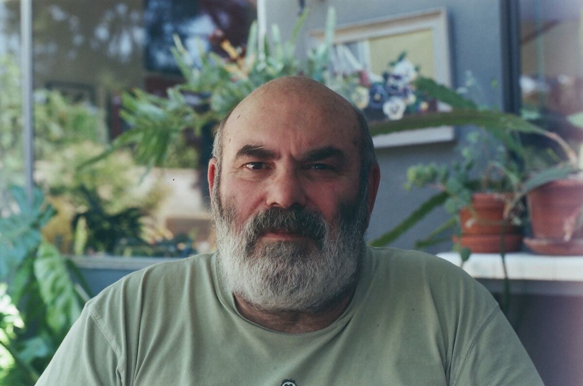 The bearded Luciano Perna in a light green T-shirt sits before plants.