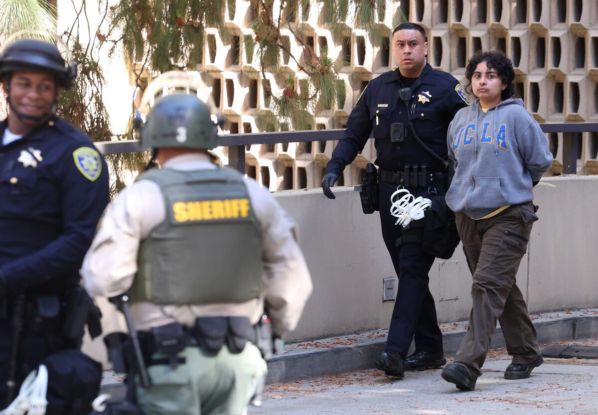 A police officer escorts a person wearing a UCLA sweatshirt down a pathway.