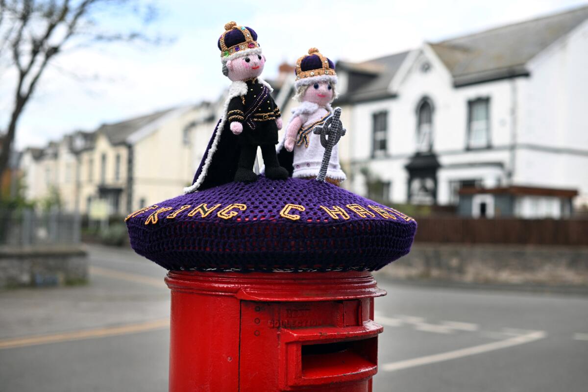 Two doll figures depicting a king and a queen, wearing crowns, adorn the top of a purple knitted cover over a red mailbox 