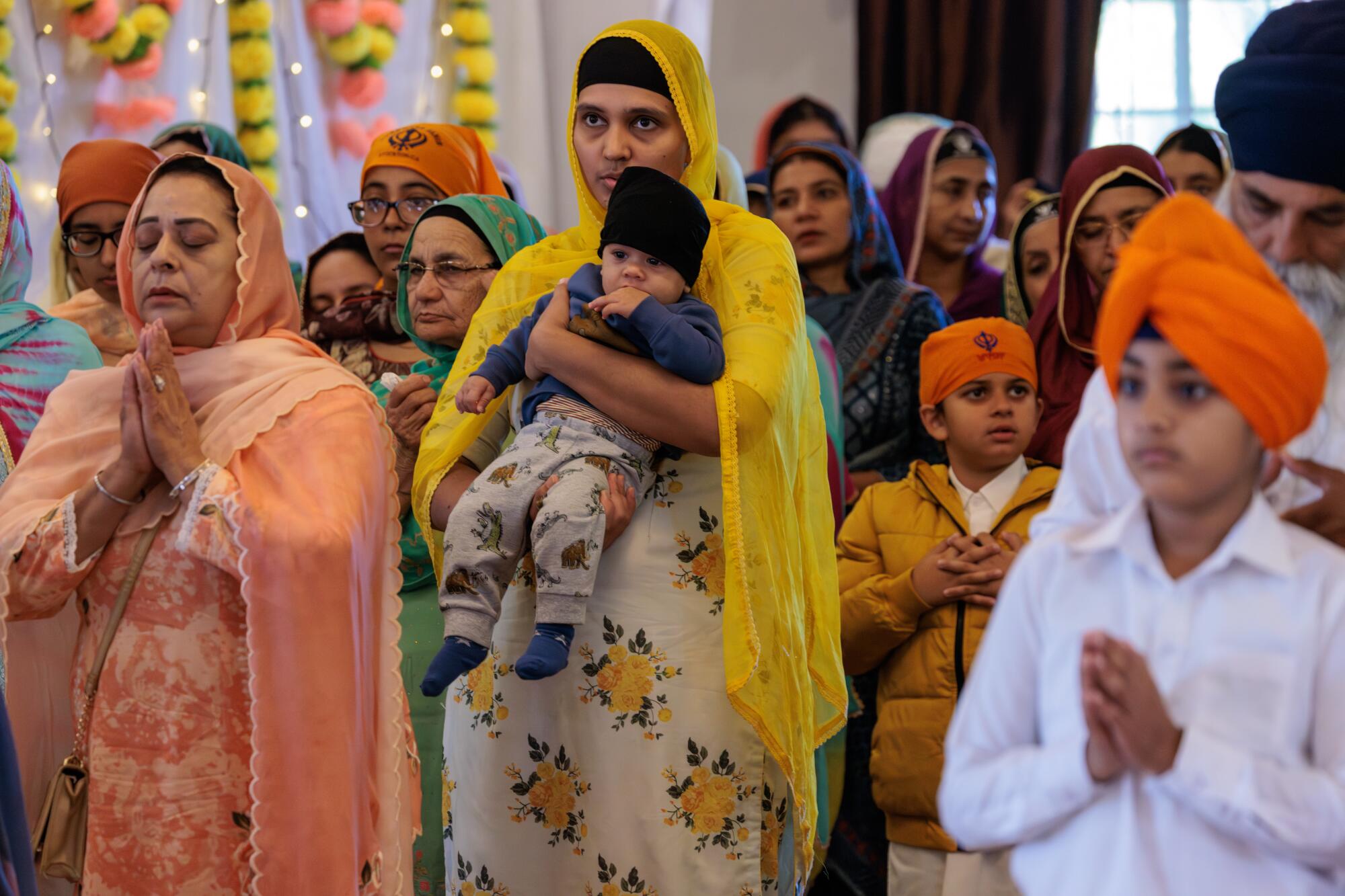 A woman in a yellow head covering, holding a young child, is flanked by another woman and orange-turbaned children