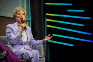 A woman in a lavender suit sits in a chair while speaking into a microphone.