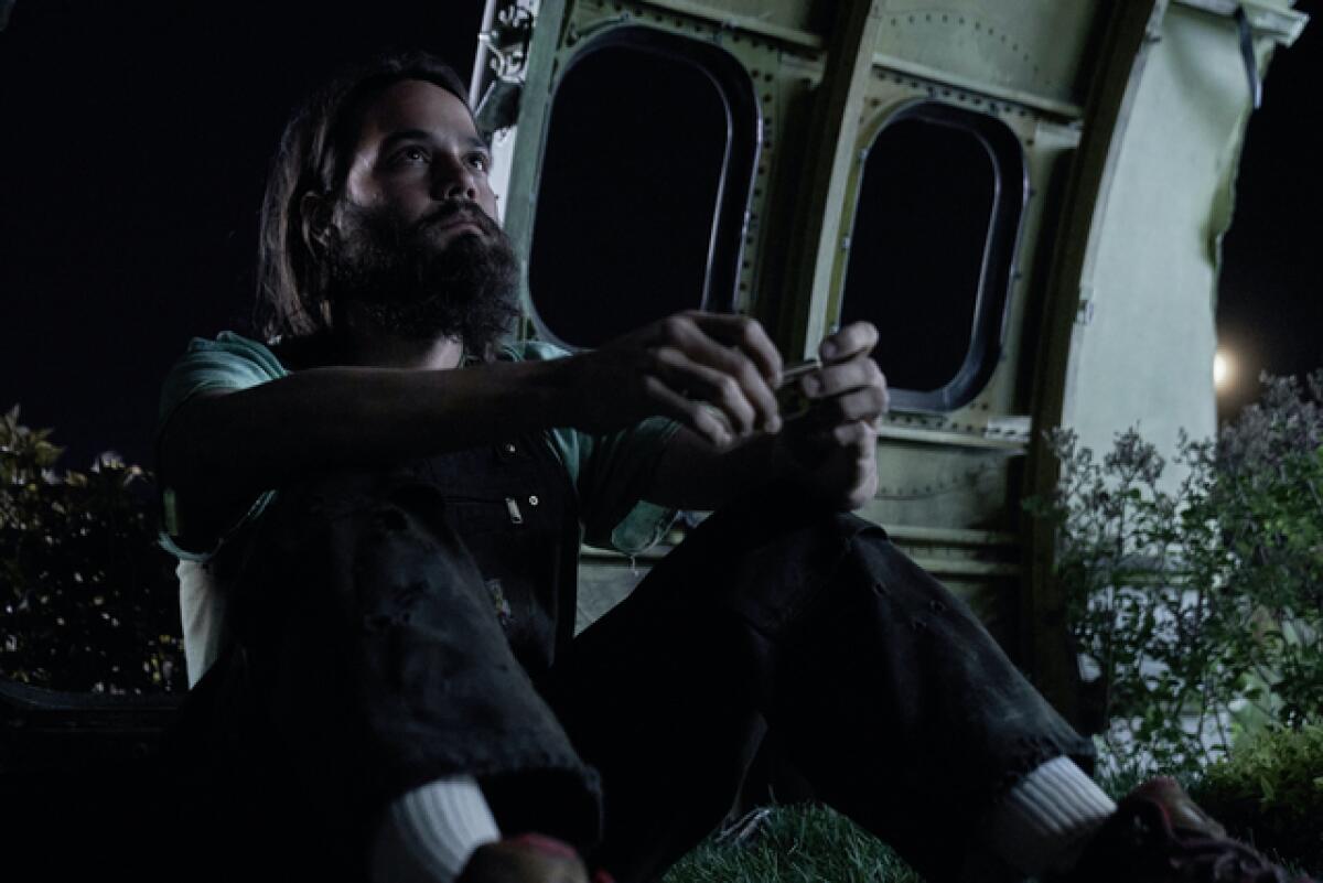 A bearded man sits against a piece of a burnt-out airplane fuselage