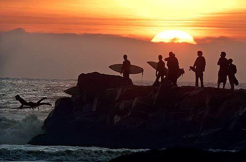 As the sun sets on a day of big waves, a young surfer leaps off a jetty in Oceanside.