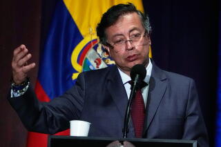 A man with dark hair, in glasses, dark suit and red tie, gestures with one hand while speaking at a lectern