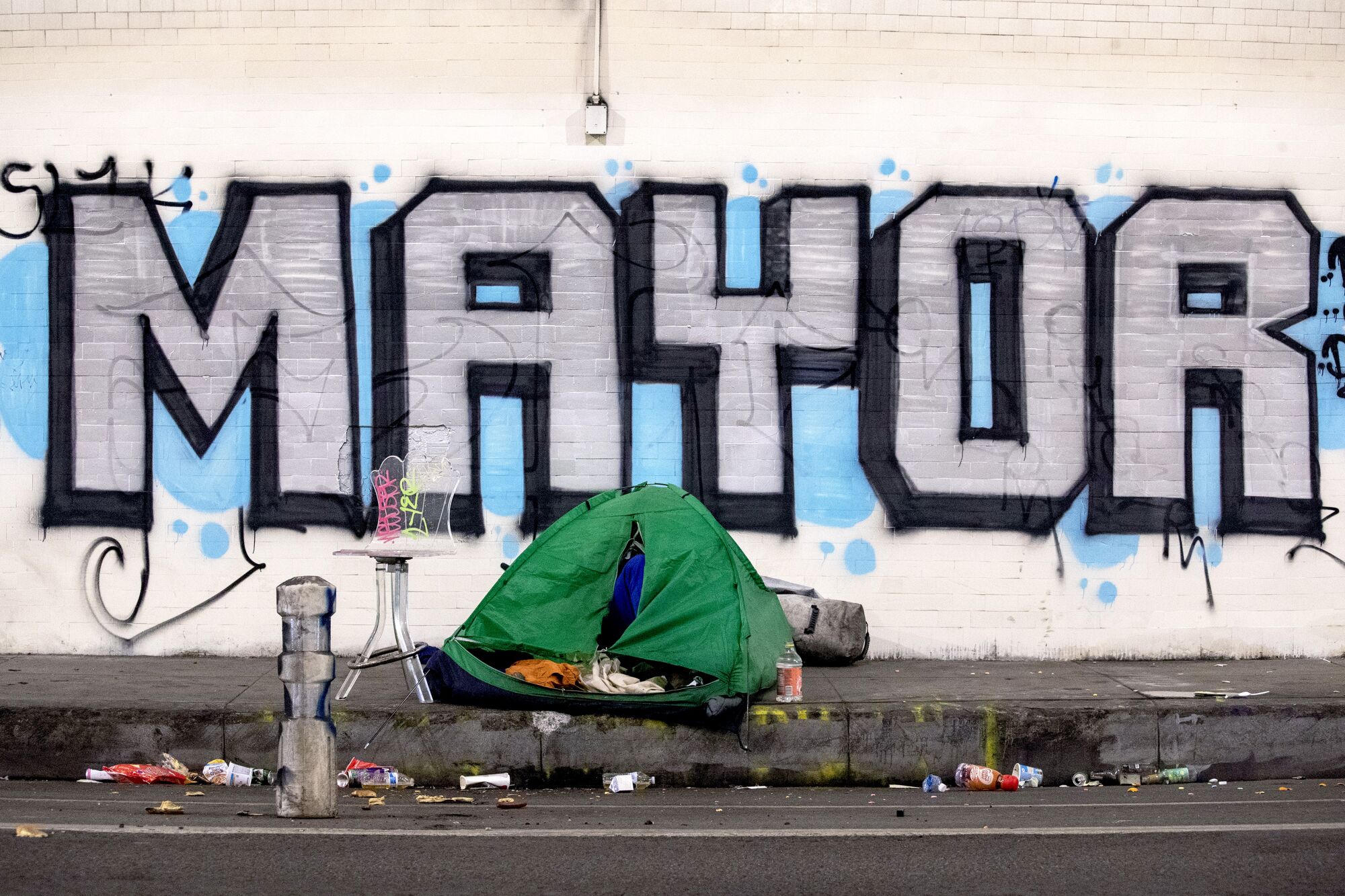 A tent, surrounded by litter, sits on the sidewalk. A large spray-painted sign behind reads "Mayor."