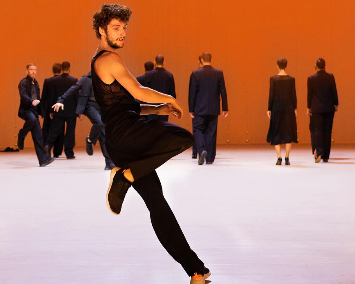 A man in black dances on one leg in front of a group of men and women in black, most with their backs to him.