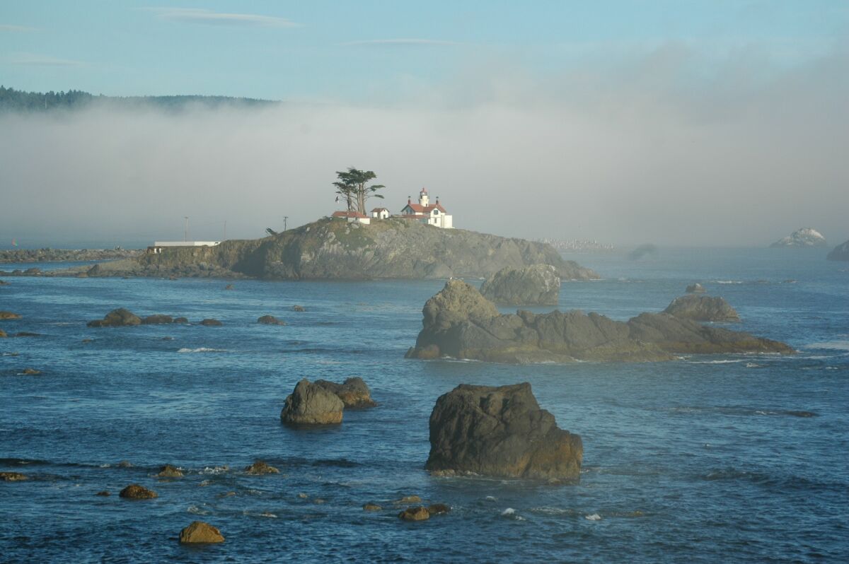 A lighthouse sits on a rock formation jutting out of the ocean, with fog behind.