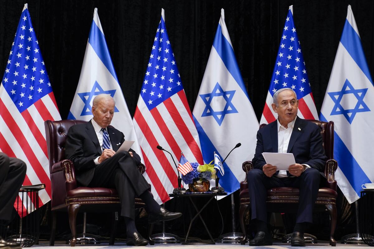 Two men sit in chairs with U.S. and Israeli flags behind them. 
