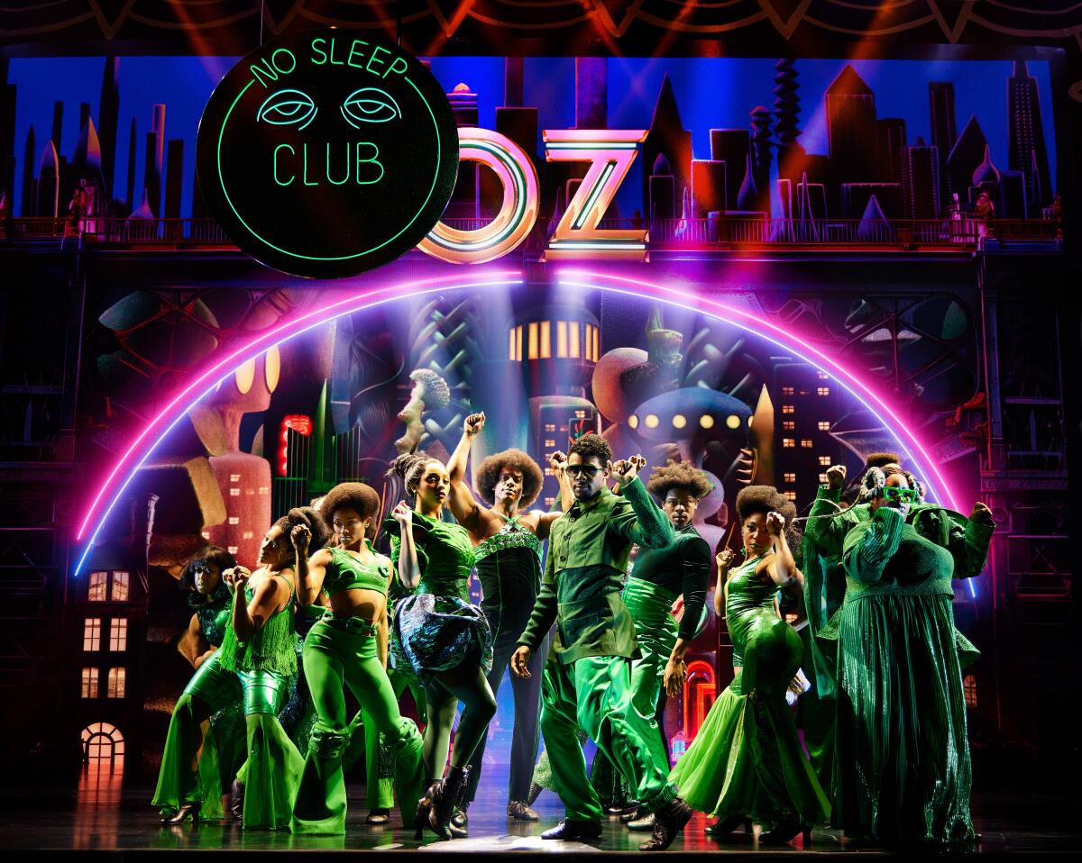 Actors dressed in green under a neon arch with the word Oz at its apex