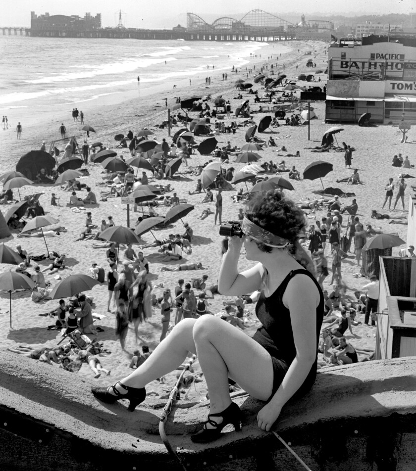 A woman in swimsuit and scarf around her head looks through binoculars. Below her is a crowded beach.