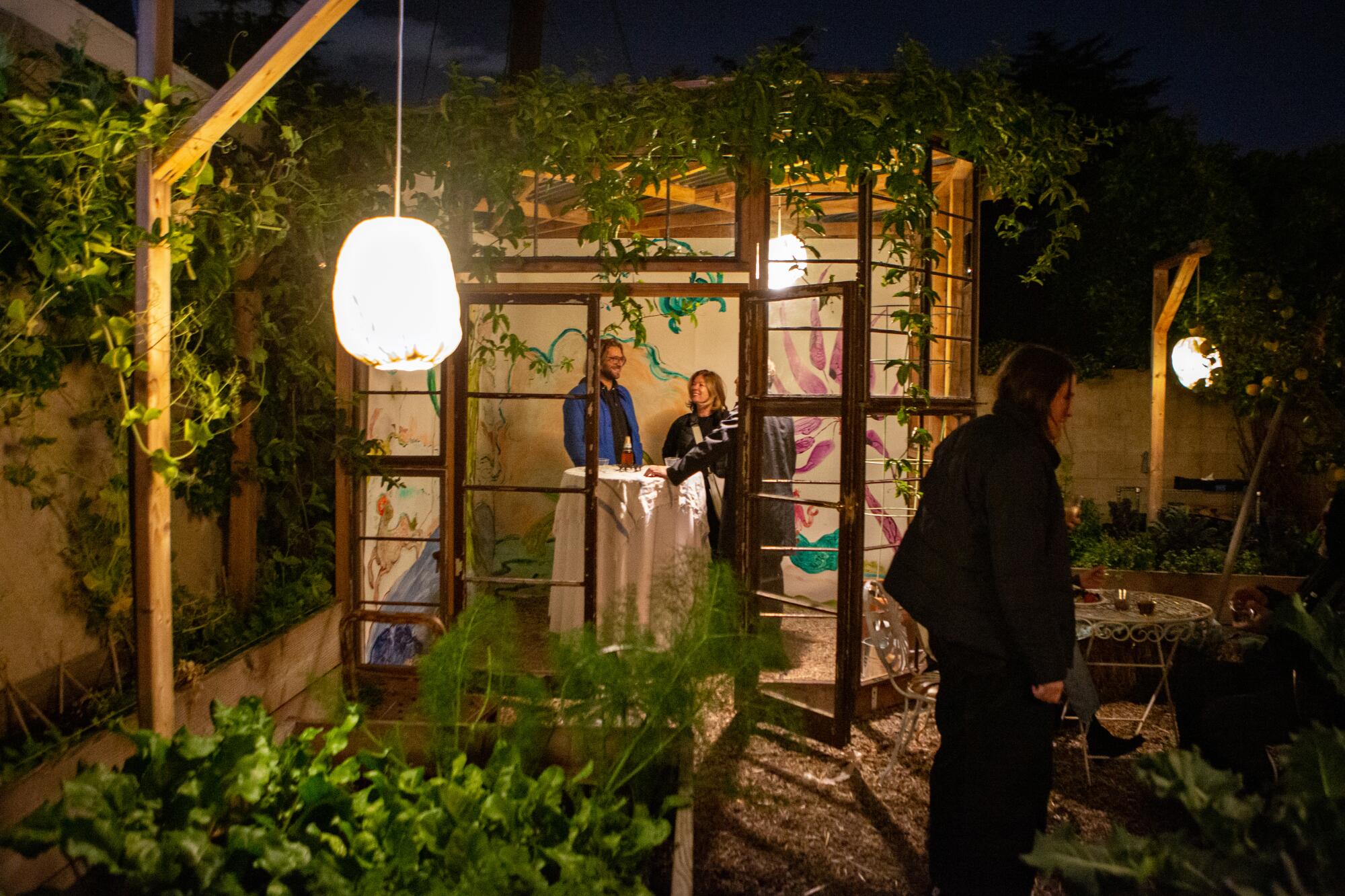 People stand in a windowed green-house gazebo filled with art on its walls 