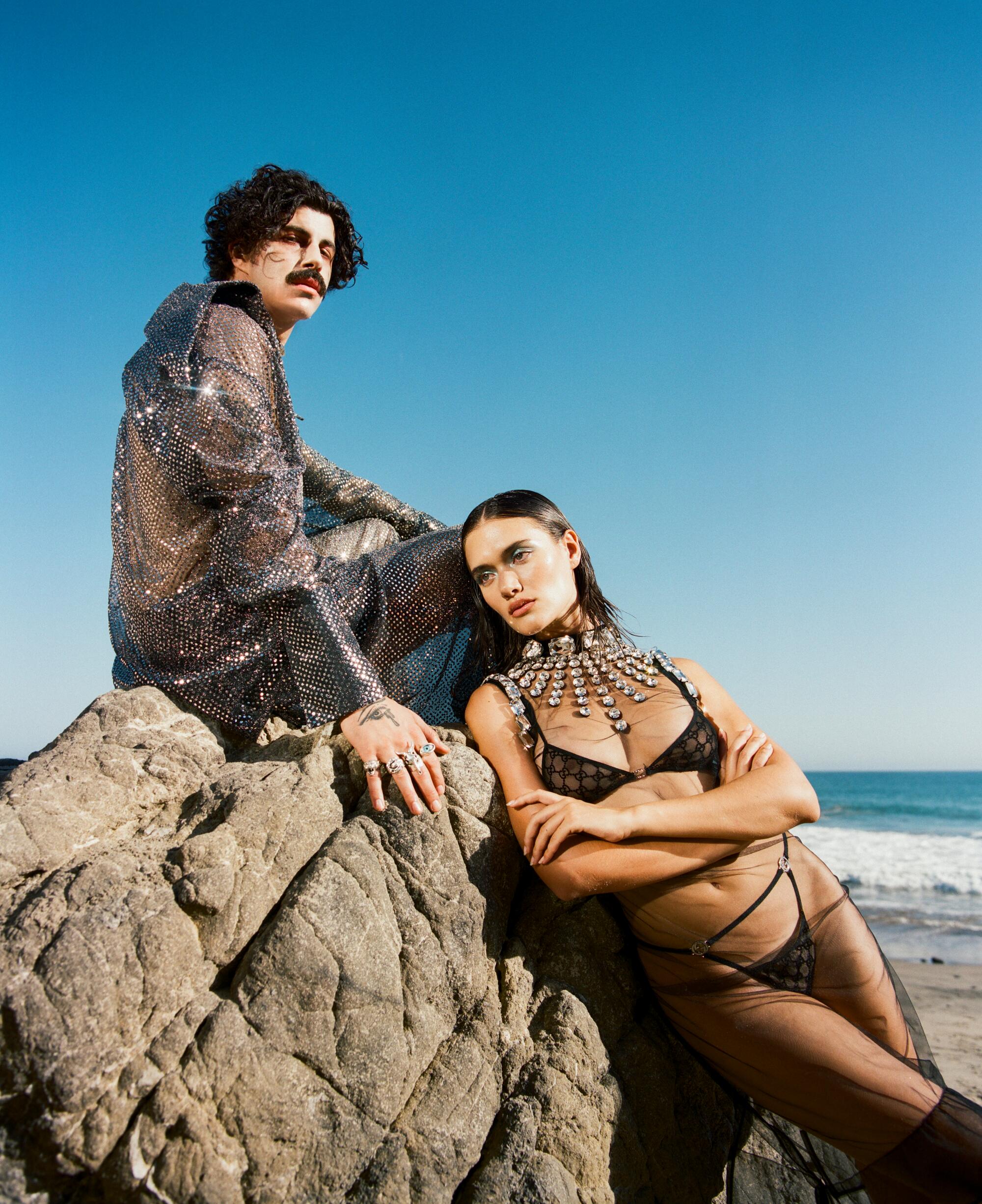 Two people sit on a rock on the beach. One wears a glittery outfit, the other wears black lingerie.