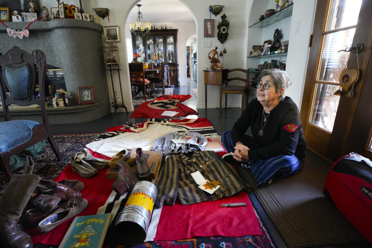An older woman sits cross-legged on the floor of a living room, surrounded by Holocaust and World War II artifacts.
