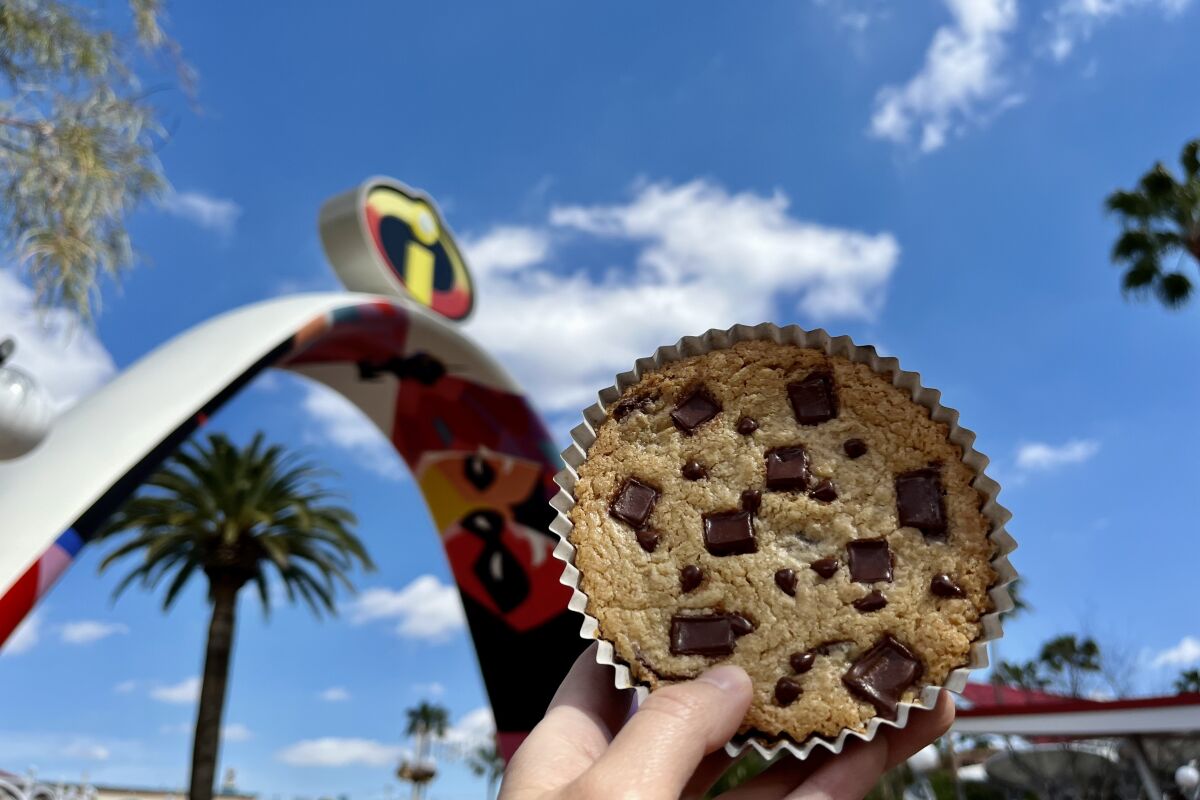A hand holds up a large chocolate chip cookie in front of the IncrediCoaster in Disney California Adventure
