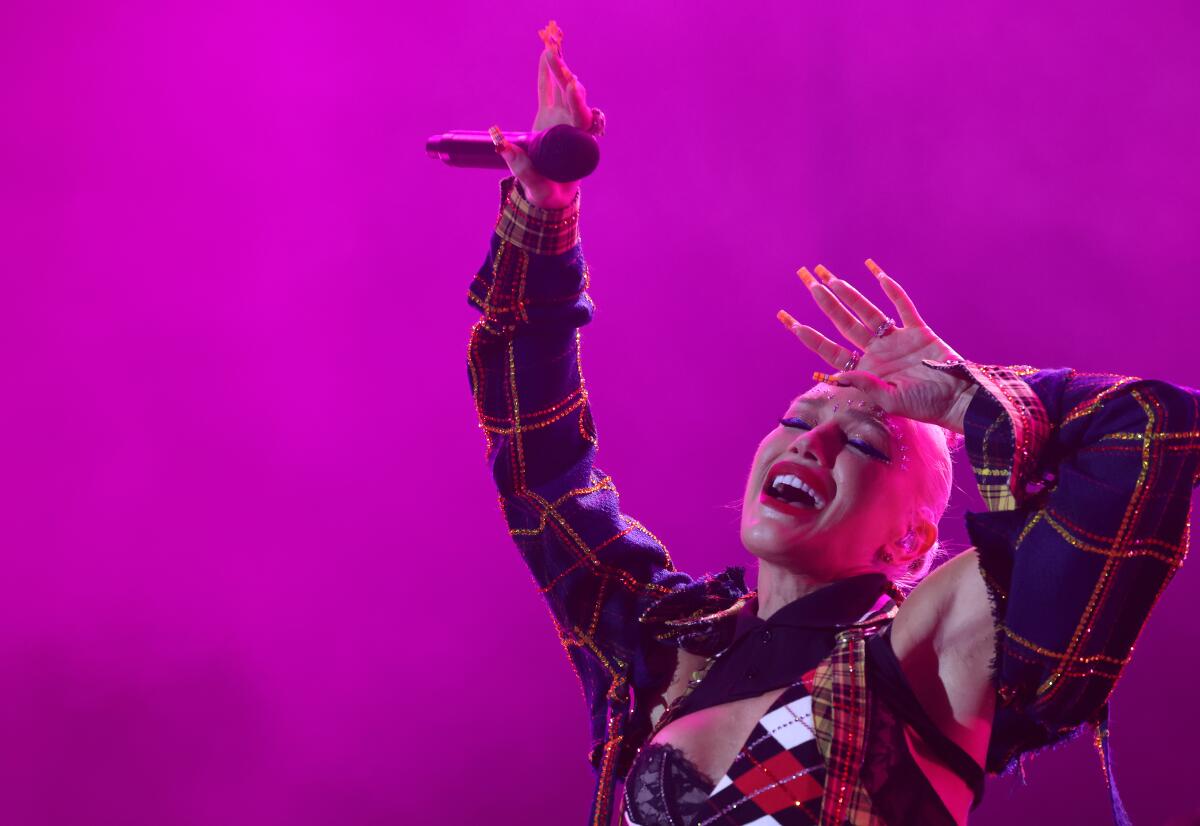 Gwen Stefani holds up a microphone with one hand and holds her other hand against her forehead.