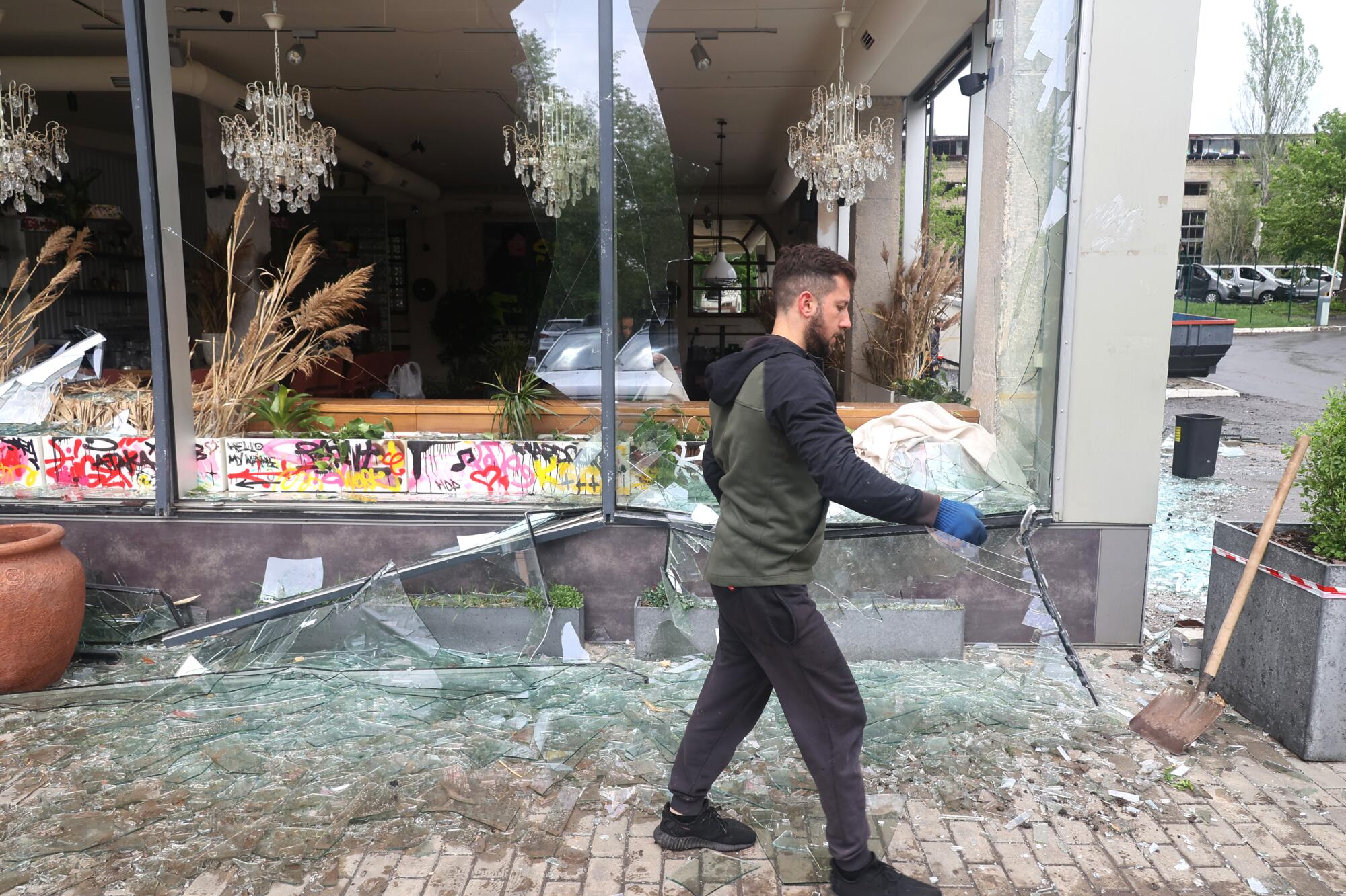 A man carries broken glass in front of a shop with shattered windows.