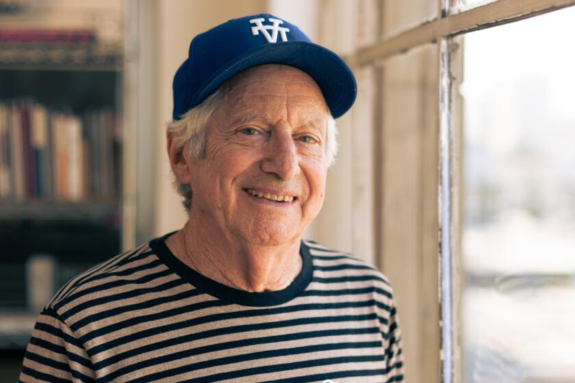 Robbie Conal stands by a window in a striped black and white shirt and a baseball cap with the letters "LA" upside down