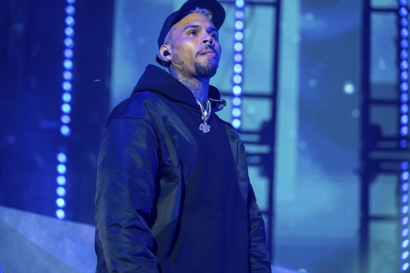Chris Brown wears a black cap and dark jacket while standing on a blue-lit stage