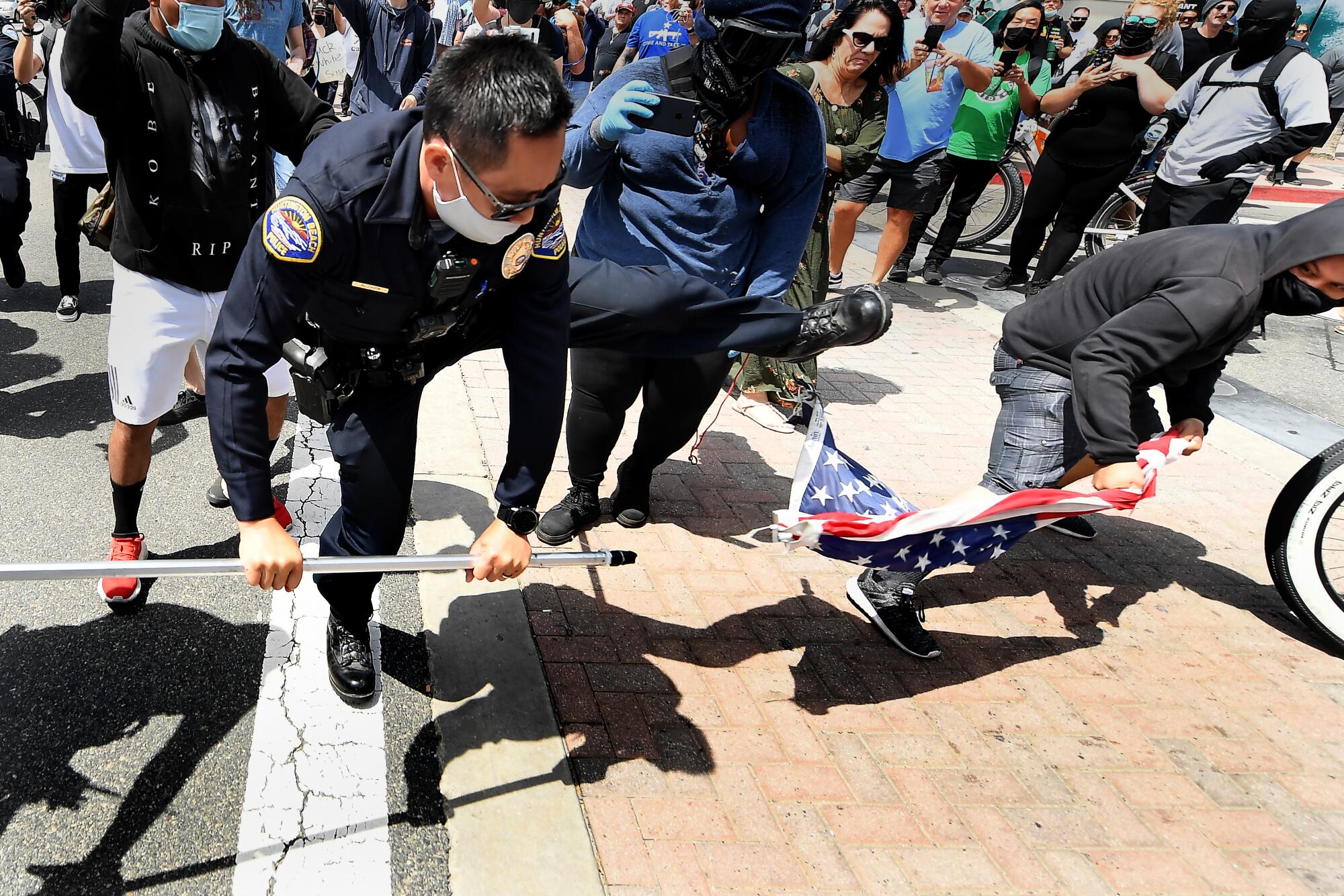 A crowd watches a police officer trying to intervene as a man takes an American flag