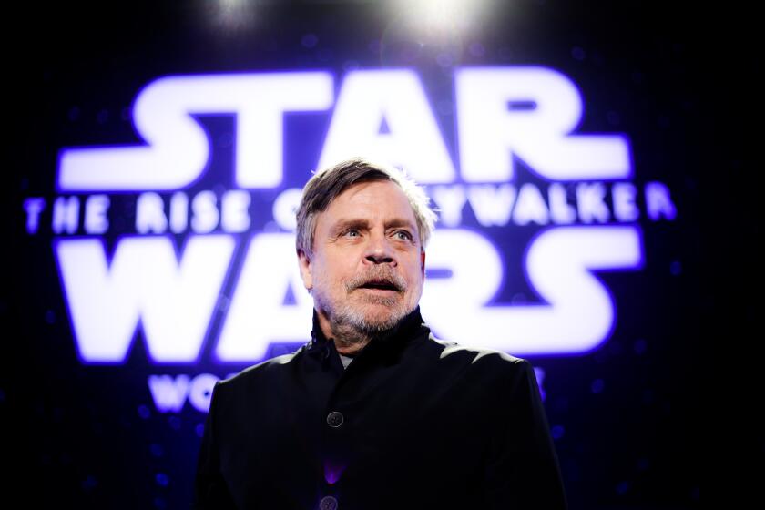 Mark Hamil is standing with his mouth agape, looking out toward audience with Star Wars written on a screen behind him