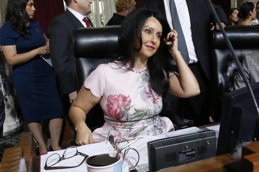 Nury Martinez, in a pink dress, is shown sitting in the City Council chambers before a wooden plaque that bears her name