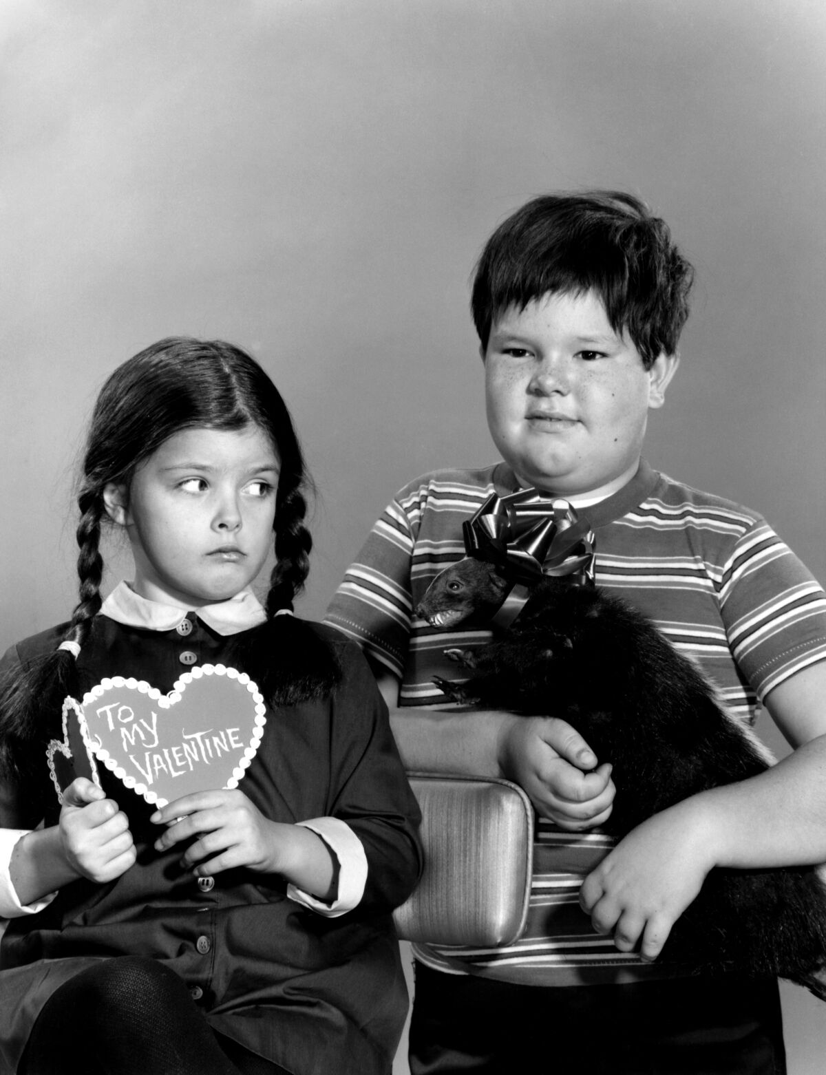 A black-and-white image of a little girl with long braids holding a valentine next to a little boy holding a stuffed animal