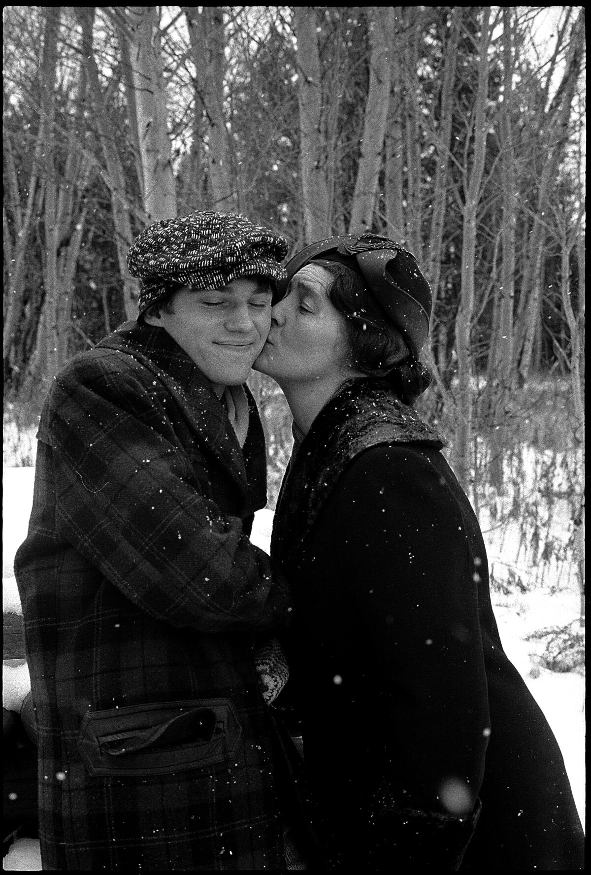 A black-and-white photo of a woman kissing a man's cheek in front of bare trees
