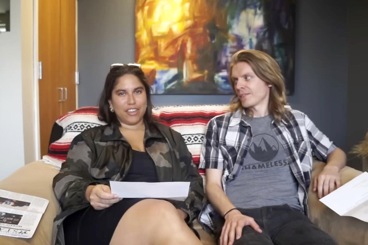 Two people sit on a small sofa. One is holding a piece of paper, speaking, and the other one looking at her. 