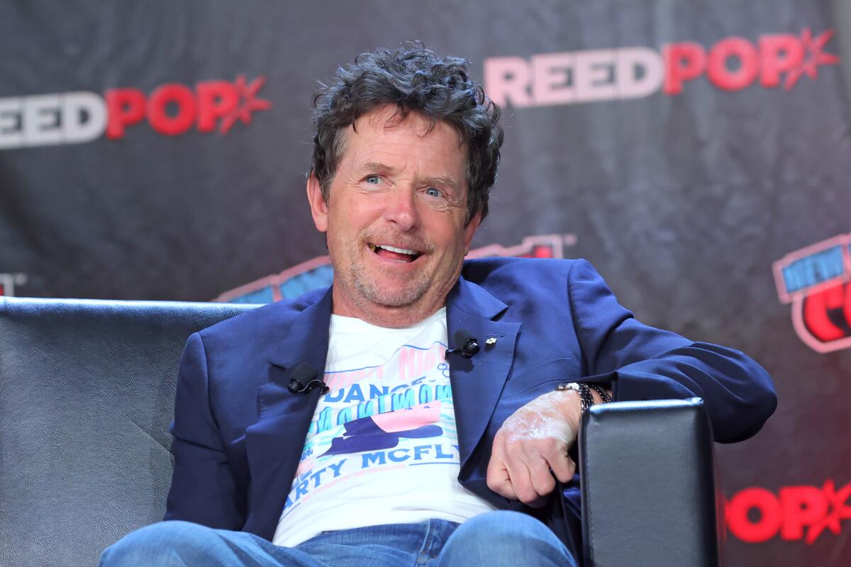 Michael J. Fox sits on black couch and smiles with his mouth open, wearing a blue blazer and white printed T-shirt