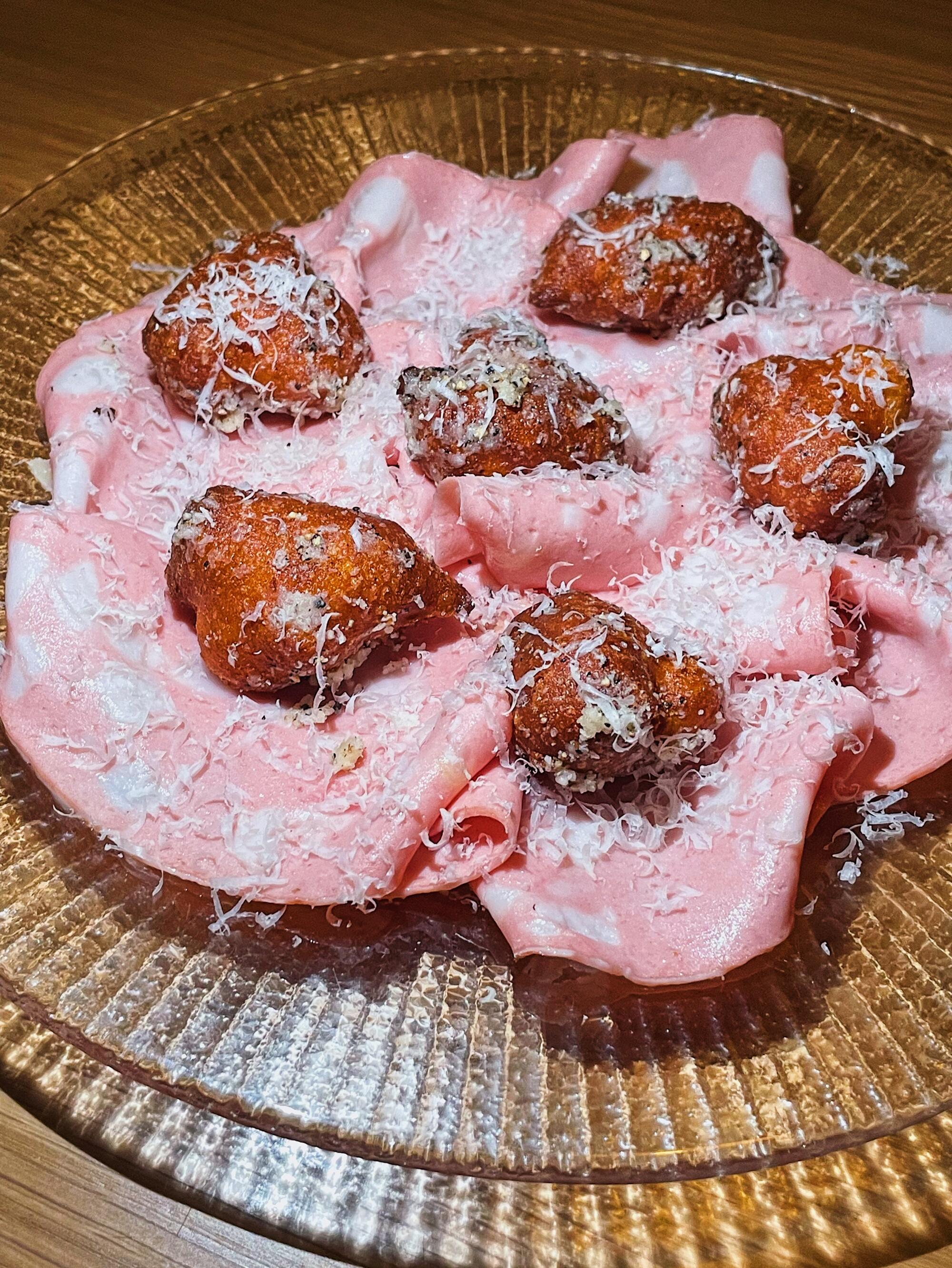 A vertical, closeup photo of mortadella on a glass plate. Atop the meat are six small savory doughnuts and grated cheese.