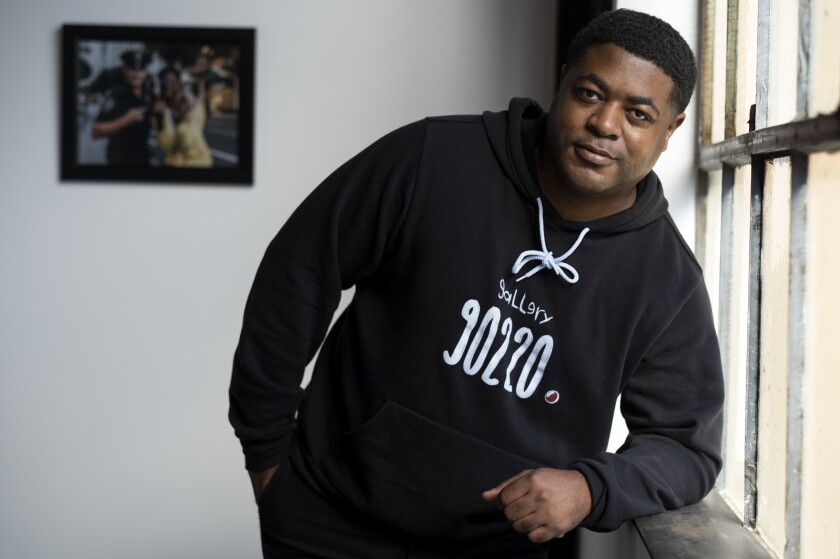 A man wearing a hoodie that reads "Gallery 90220" leans on a window sill.