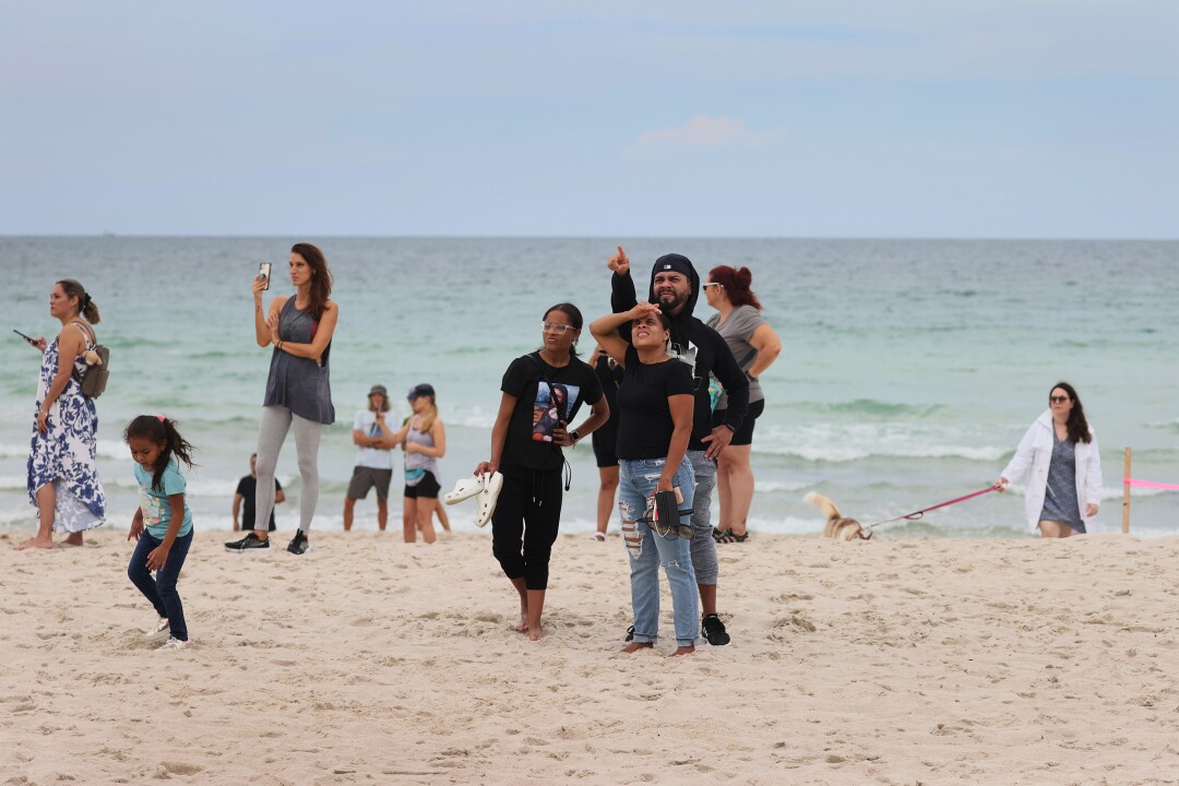 Children and adults stand on the sand, some recording with cellphones. One man points as his family gazes upward.