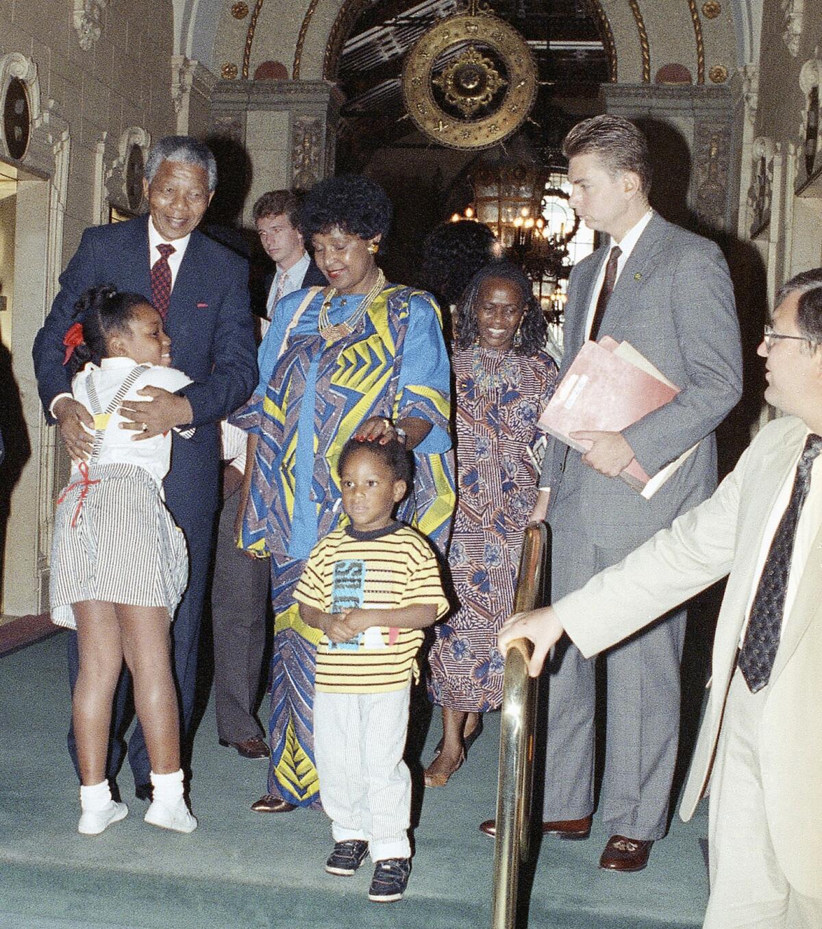 A young girl hugs a man in a suit and tie. Beside him stands a woman, who has her hand on the head of a young boy.