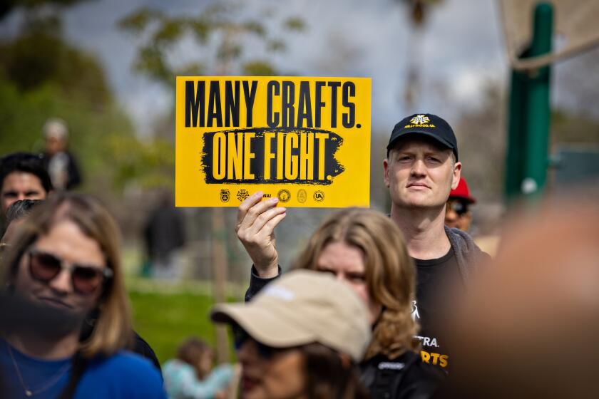 A man in a crowd wearing an IATSE hat and holding a yellow sign that reads, "Many crafts. One fight."