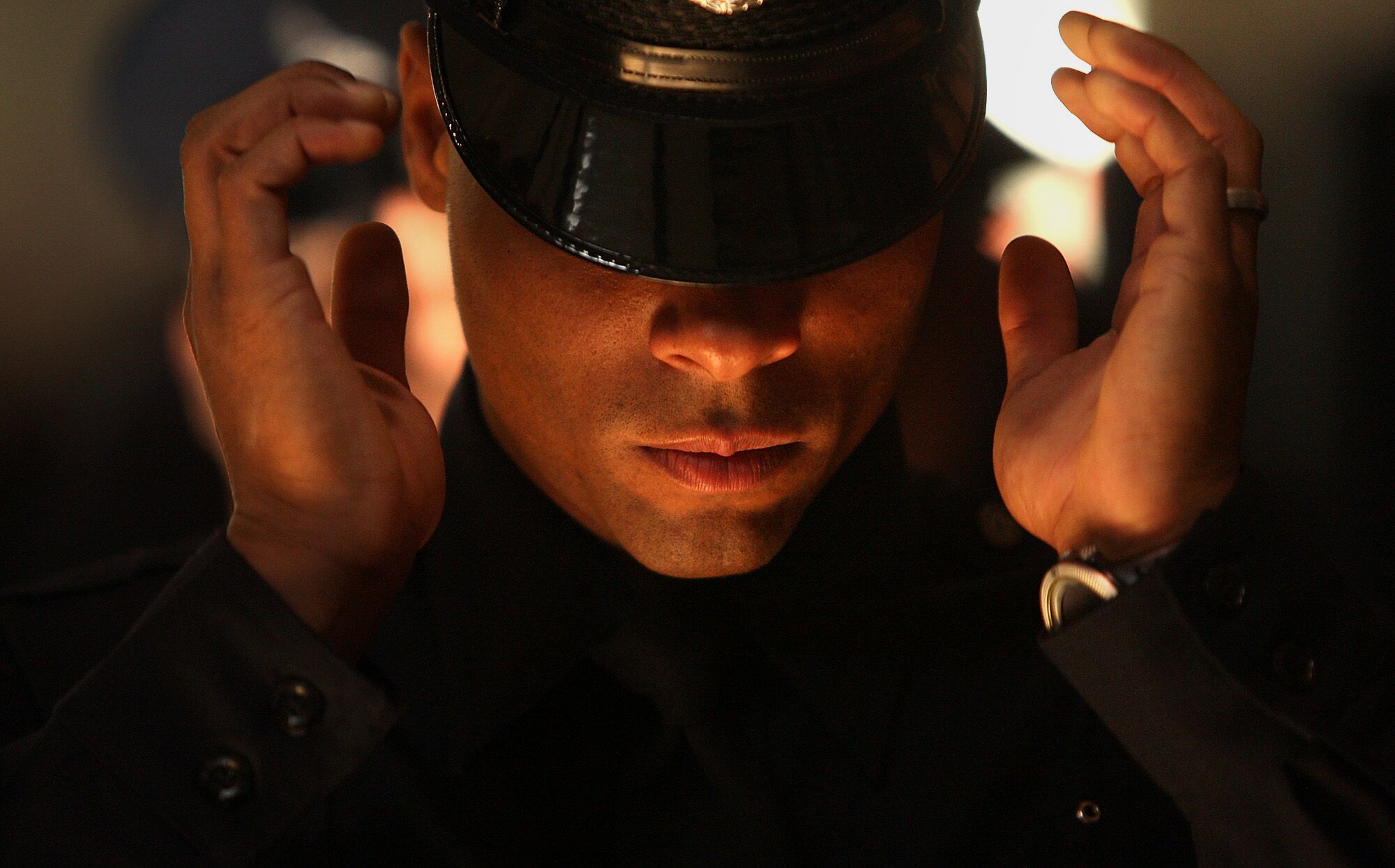 In a head-and-shoulders frame a police officer reaches with his hands to adjust his police cap, which is shielding his eyes.