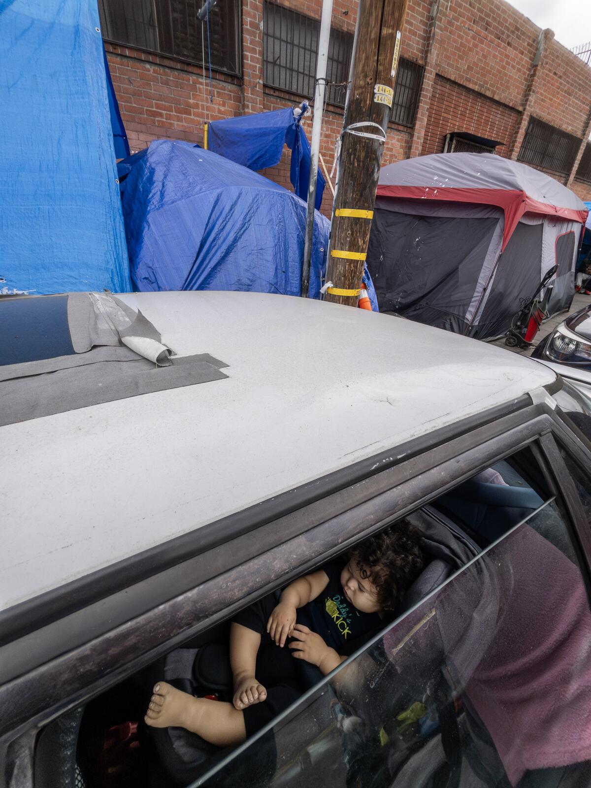 A young child is in a car with a window partly rolled down, with tents nearby.