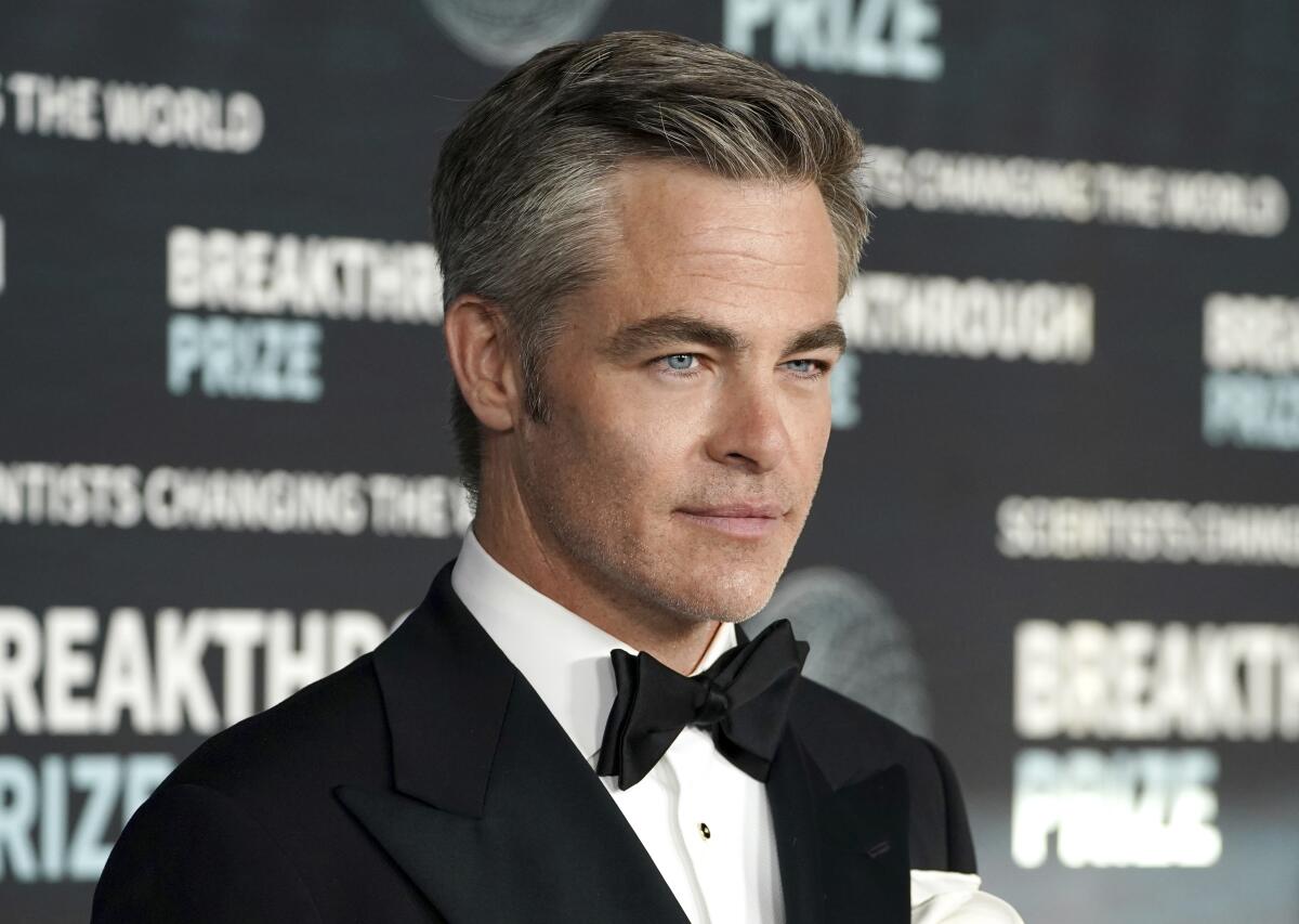 A closeup of Chris Pine's face showing the top of his tuxedo and black tie in front of a backdrop