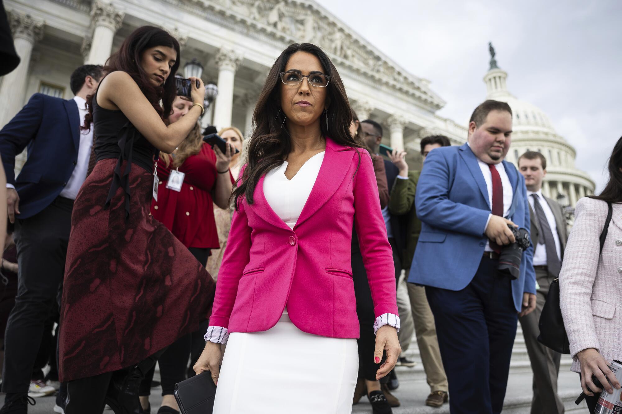 A woman with dark hair, in a white dress and deep pink jacket, stands near other people outside the Capitol