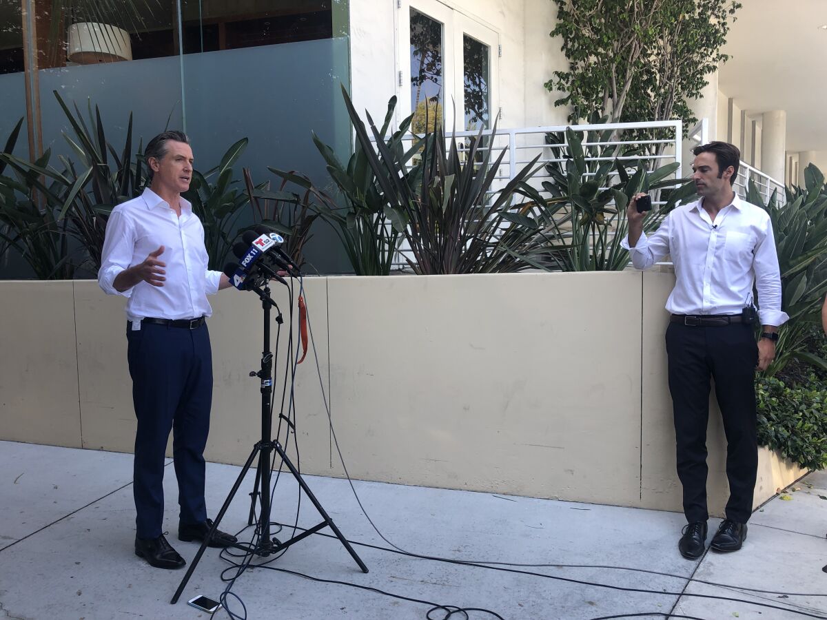 A man in dark pants and white button-down stands on a sidewalk and talks into microphones.